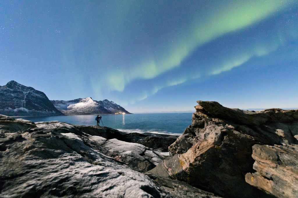 Photographer under the stars and Northern Lights (aurora borealis) surrounded by rocky peaks and icy sea, Tungeneset, Senja, Troms, Norway, Scandinavia, Europe