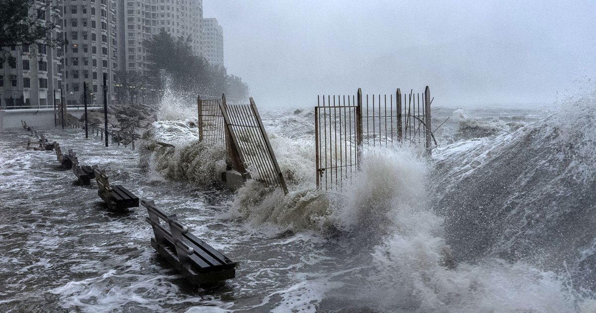 Strong waves crash against secured fences and benches along a city waterfront, with high-rise buildings visible in the background amidst rough, stormy conditions caused by tropical cyclone Hong Kong November 2025.